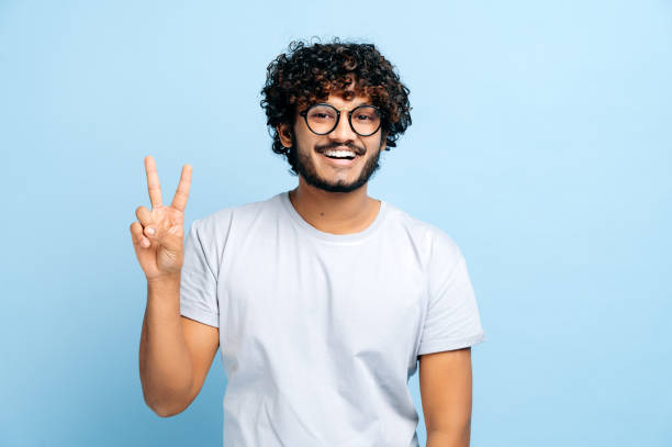 istock Charismatic handsome positive indian or arabic guy with glasses, freelancer or creative designer, stands on a blue isolated background, shows a PEACE sign, looks at the camera, smiles friendly 1392990656