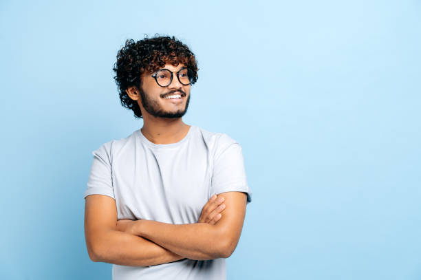 attraente ragazzo positivo indiano o arabo dai capelli ricci con gli occhiali, che indossa una maglietta di base, studente o libero professionista, in piedi su uno sfondo blu isolato, con le braccia incrociate, guarda di lato, sorride - look right foto e immagini stock