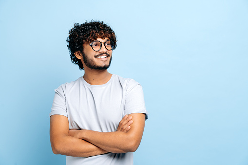 Attractive positive Indian or Arabic curly haired guy with glasses, wearing a basic t-shirt, student or freelancer, standing over isolated blue background, with arms crossed, looks to the side, smiles