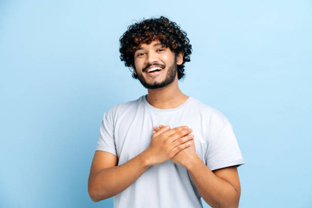 un geste de sincérité et de confiance. beau gars indien ou arabe candide avec les mains sur le cœur et un geste reconnaissant, sourire sur le visage, regarde la caméra, se tient sur un fond bleu isolé. concept paisible - smile sign photos et images de collection