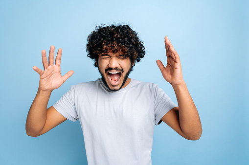 Annoyed curly-haired indian or arabian guy in a t-shirt, shouting loudly with his eyes closed, gesturing with his hands, standing on isolated blue background. Emotion of annoy, anger and disagreement