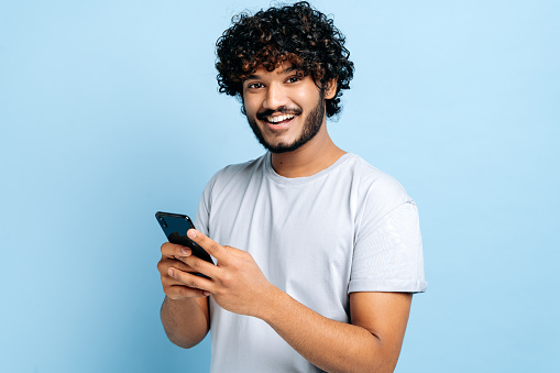 Positive indian or arabian guy, wearing a basic t-shirt, using his smartphone, typing a message, browsing the internet, social media, stands on isolated blue background, looking at camera, smiling