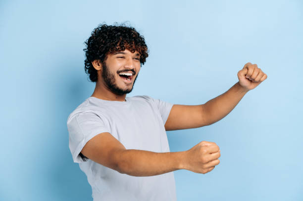 Joyful attractive curly haired indian or arabian guy, wearing casual t-shirt, holding in hands driving invisible car, imaginary steering wheel, stands on isolated blue background, smiling stock photo