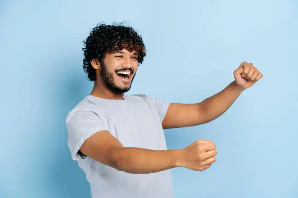 Photo of Joyful attractive curly haired indian or arabian guy, wearing casual t-shirt, holding in hands driving invisible car, imaginary steering wheel, stands on isolated blue background, smiling