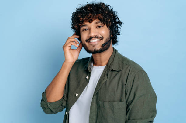 portrait d’un jeune homme adulte indien ou arabe positif amical, centre d’appels, employé de la hotline avec casque et microphone, se tient sur fond bleu isolé, regarde la caméra, souriant - indian ethnicity audio photos et images de collection