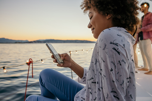Woman with curly hair and brown skin smiling while looking at her smartphone, sitting on boat deck in the evening during sunset, friends standing in background, focus on foreground, view from the side