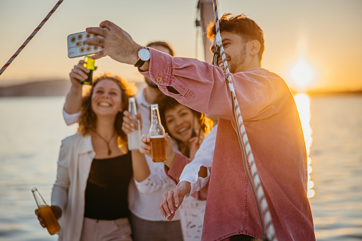 Man with short brown hair and beard taking a selfie with his friends, holding his smartphone up in the air, standing on boat deck of a sailboat during sunset, everyone holding drinks and smiling in the camera