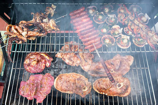 Mixed Seafood And Meat Cooking On A Barbecue Including Octopus, Prawns, Sausages And Pork