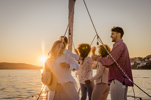 Group of friends, two men and three women, having a party on a sailboat, dancing to music while standing on front boat deck, sea with island and beautiful sunset sky in the background