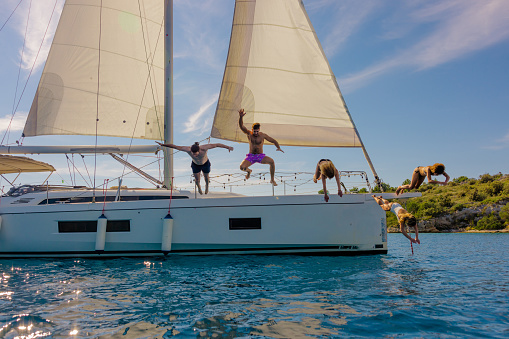 Group of friends, two men and three women, enjoying a warm summer day, wearing swimwear, jumping from boat deck of a white sailboat into the sea, front view, island and clear sky in background