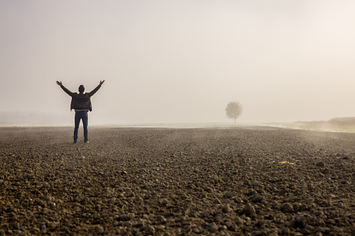 Male farmer standing with arms outstretched on plowed agricultural field covered with fog