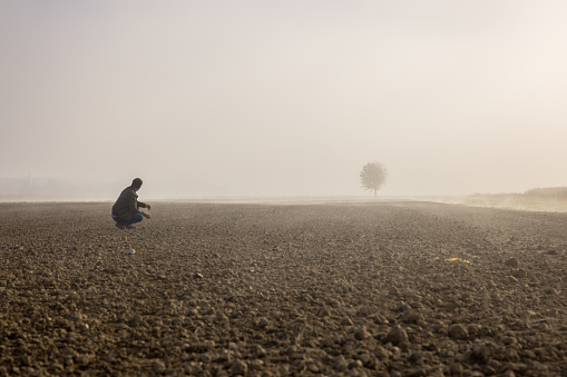 Male farmer examining soil at plowed agricultural field covered with fog