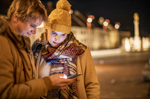 Smiling young couple using smart phone at illuminated city during Christmas night in winter