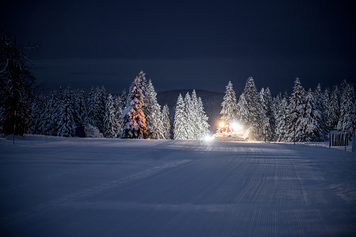 Illuminated snow groomer against forest on snowy landscape during winter at night
