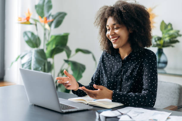 Successful positive young adult woman african woman freelancer, manager, CEO, sitting in office at laptop, talking on video call with client or employees, discussing business strategy, gesturing,smile stock photo