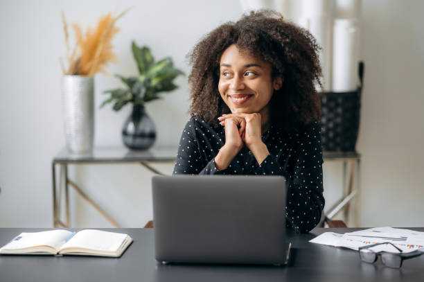 istock Satisfied dark-skinned curly-haired young woman, office worker, manager or financier, sits at a desk in modern office, rests from work, takes break,  looks away, smiles, dreaming of rest 1392996234