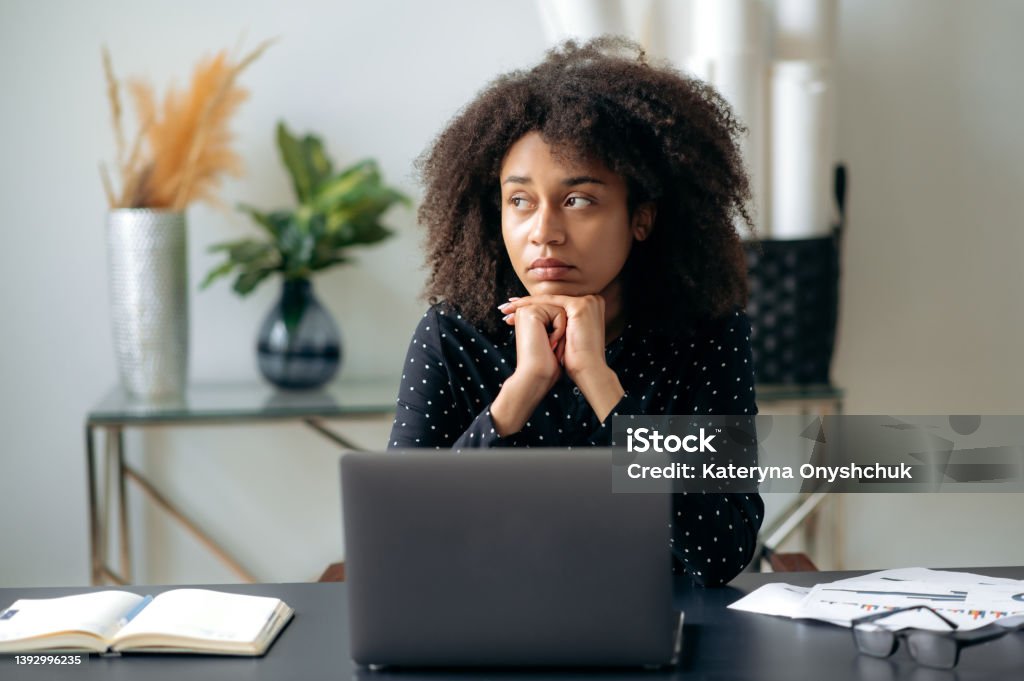Tired african american curly haired woman, business lady, director, manager, sits in front of a laptop at her desk, rests from computer work, looks to the side, dreams of rest or vacation Disappointment Stock Photo