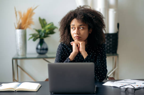 Tired african american curly haired woman, business lady, director, manager, sits in front of a laptop at her desk, rests from computer work, looks to the side, dreams of rest or vacation stock photo