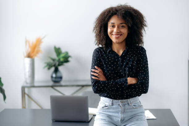 chica afroamericana encantadora y positiva. retrato de mujer exitosa, freelancer, gerente, elegantemente vestida, de pie cerca del escritorio en la oficina moderna, mira a la cámara, con los brazos cruzados, sonríe amigablemente - aprendiz fotografías e imágenes de stock