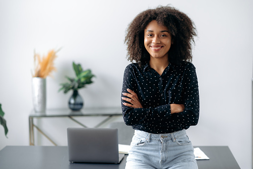 Positive lovely african american girl. Portrait of successful woman, freelancer, manager, stylishly dressed, stand near desktop in modern office, look at the camera, with arms crossed, smiles friendly