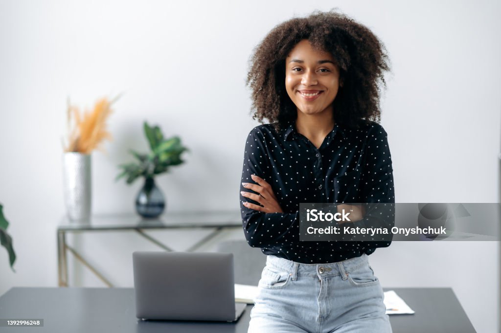 Positive lovely african american girl. Portrait of successful woman, freelancer, manager, stylishly dressed, stand near desktop in modern office, look at the camera, with arms crossed, smiles friendly Trainee Stock Photo