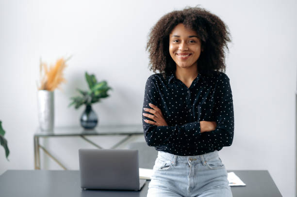 Positive lovely african american girl. Portrait of successful woman, freelancer, manager, stylishly dressed, stand near desktop in modern office, look at the camera, with arms crossed, smiles friendly stock photo