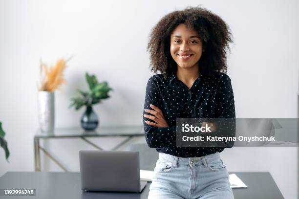 Positive Lovely African American Girl Portrait Of Successful Woman Freelancer Manager Stylishly Dressed Stand Near Desktop In Modern Office Look At The Camera With Arms Crossed Smiles Friendly Stock Photo - Download Image Now