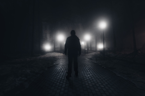 Sad man alone walking along the alley in night foggy park. Back view.