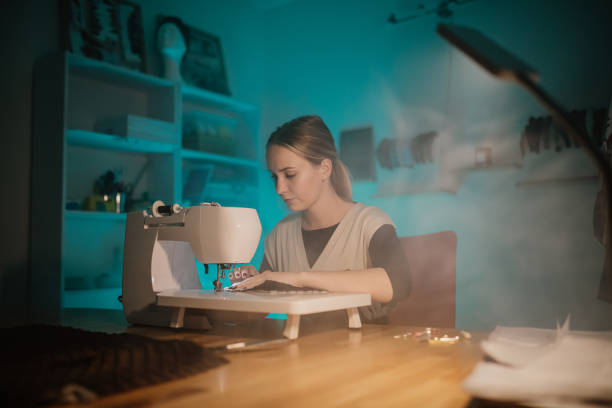 Young female fashion designer using sewing machine to stitch fabric on table in modern boutique