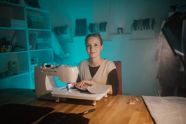 Smiling young female fashion designer using sewing machine to stitch fabric on table in modern boutique