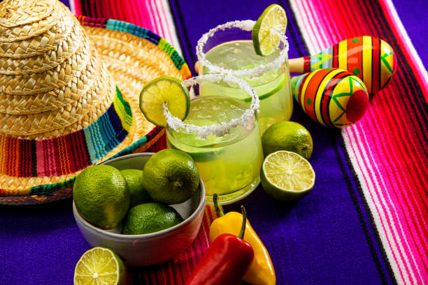 Happy Cinco de Mayo with two Margarita Glasses on a Colorful Mexican Blanket This is a photograph of two modern margarita glass with a rim of salt surrounded by fresh cut limes and chilis on a colorful striped mexican blanket next to a sombrero cinco de mayo stock pictures, royalty-free photos & images