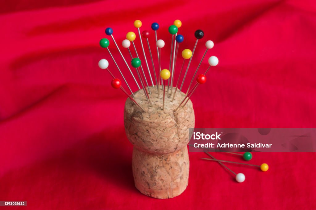 Sewing pins in cork pincushion on red fabric Multi-colored, ball head straight pins used for sewing stuck into a champagne cork pincushion, centered on a background of bright red fabric. Cork - Stopper Stock Photo