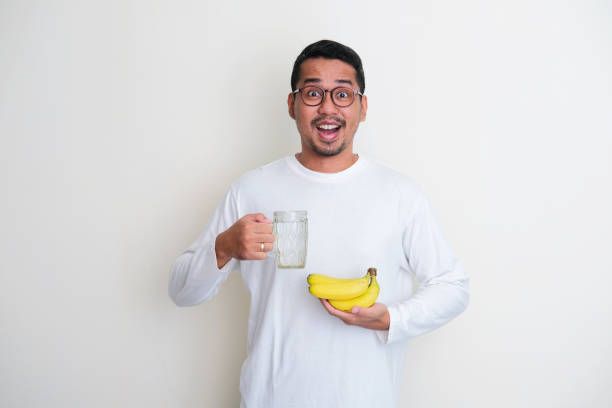 uomo asiatico adulto che mostra un'espressione wow quando tiene in mano un bicchiere d'acqua e banana - men banana holding cut out foto e immagini stock