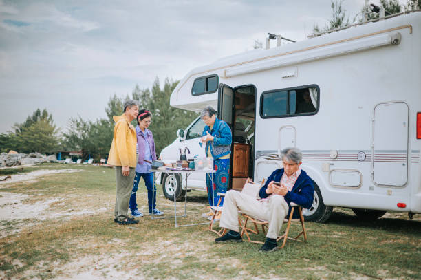 messagerie texte d’un homme âgé chinois d’origine asiatique pendant que ses amis préparaient le petit-déjeuner du matin à côté d’un camping-car garé sur la plage du parc à roulottes - motor home mobile home camping senior adult photos et images de collection