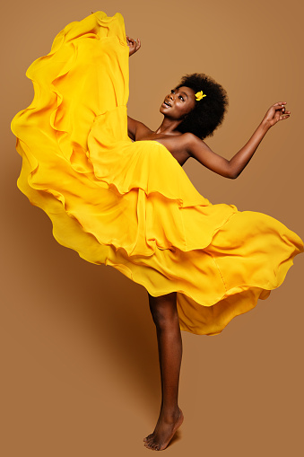 Expressive Woman dancing in Yellow Flying Dress. Happy Dark Skinned Dancer in Waving Fabric Gown. Model with Black curly Afro Hair jumping over Beige Background full length