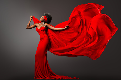 Fashion African Woman in Red Dress with Silk Scarf flying on Wind. Happy Dark Skinned Model Carefree Dancing with Fluttering Chiffon Fabric over Gray Background. Stylish Afro Female with Curly Hairstyle in Long Evening Luxury Gown with waving Cloth