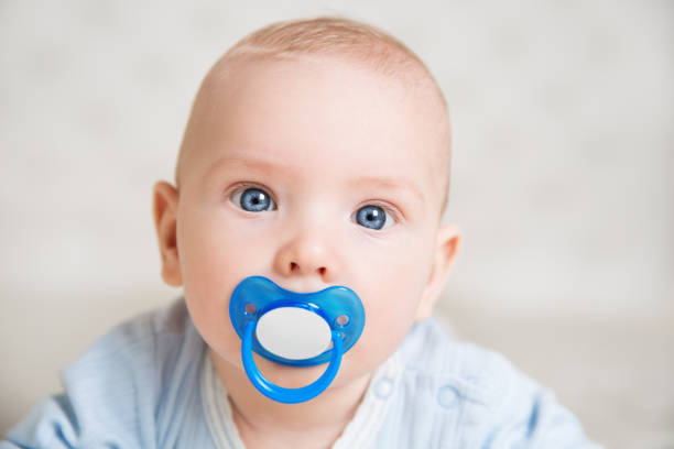 Baby sucking Pacifier looking at Camera. Child with Kids Smoother. Infant Boy Face Portrait with Dummy and Blue big Eyes in white Bed Room lying down on Stomach Baby sucking Pacifier looking at Camera. Child with Kids Smoother. Infant Boy Face Portrait with Dummy and Blue big Eyes in white Bed Room lying down on Stomach nipple stock pictures, royalty-free photos & images