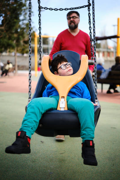 Disabled boy swinging on a special needs swing while enjoying with his father at the playground. Disabled people and parenthood concept.