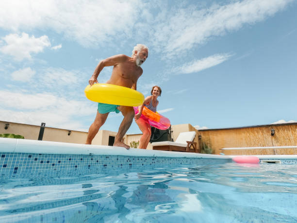 Young at heart Photo of a senior couple refreshing on a hot summer day in 
 swimming pool; they are jumping into the pool while wearing inflatable rings one piece swimsuit photos stock pictures, royalty-free photos & images