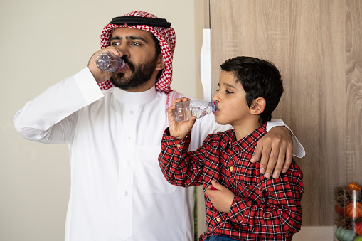 Waist-up view of Middle Eastern man in early 30s with arm around 7 year old boy, both maintaining healthy lifestyle as they drink from water bottles.