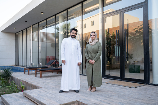 Full length view of man in dish dash and woman in abaya and hijab standing together at entrance to modern family home in Riyadh.