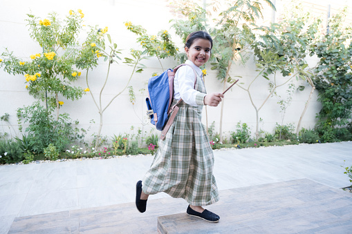 Full length side view of Middle Eastern girl in uniform with backpack smiling at camera as she runs up steps to front door after school.