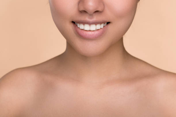 half face of asian girl. chinese young woman with perfect skin demonstrates her smile, perfect white well-groomed teeth, stands on an isolated beige background with naked shoulders. dentistry concept - half smile imagens e fotografias de stock