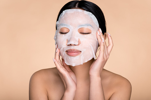 Sensual photo of beautiful young asian woman with with eyes closed, applying cotton facial moisturizing mask on face, takes care of skin, prevents wrinkles, stands on isolated beige background