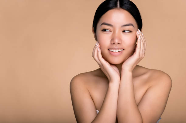 beautiful chinese brunette young woman, with perfect skin and naked shoulders, posing with hands near face at isolated beige background, looking away, smiling pleasant. beauty, care and youth concept - women tan perfection naked imagens e fotografias de stock