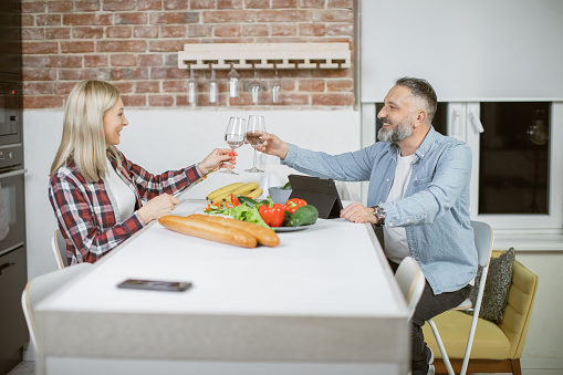 Happy mature married couple sitting at kitchen table and toasting with glasses of water. Fresh organic vegetables prepared for dish. Healthy eating and lifestyles concept.