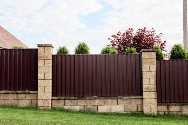 Photo of Brown metal profile fence with block posts. Incline construction. Corrugated surface. Security. Private property fencing. Opaque hedge. Outdoor house exterior. Side view. Urban or industrial style