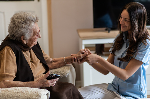 District nurse is comforting old lady during home health care visit. They are sitting in the living room, having  a friendly conversation. Senior woman is scared because of Corona virus, and  the nurse is doing the best she can to help her.