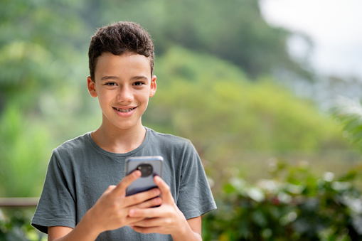 Portrait of a young teenage boy with braces smiling while standing outside with a smart phone in summer