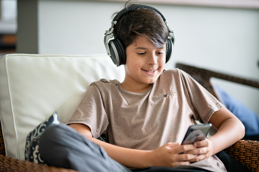 Smiling teenage boy wearing headphones and watching something on a smart phone while sitting in his living room at home